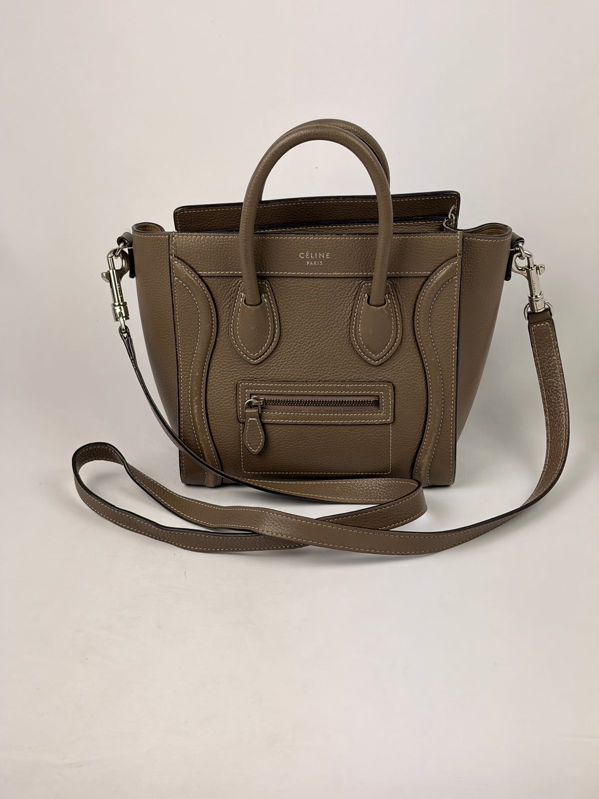 A taupe brown pebbled leather handbag with dual top handles, a front zip pocket, and a matching shoulder strap, photographed against a neutral off-white background.