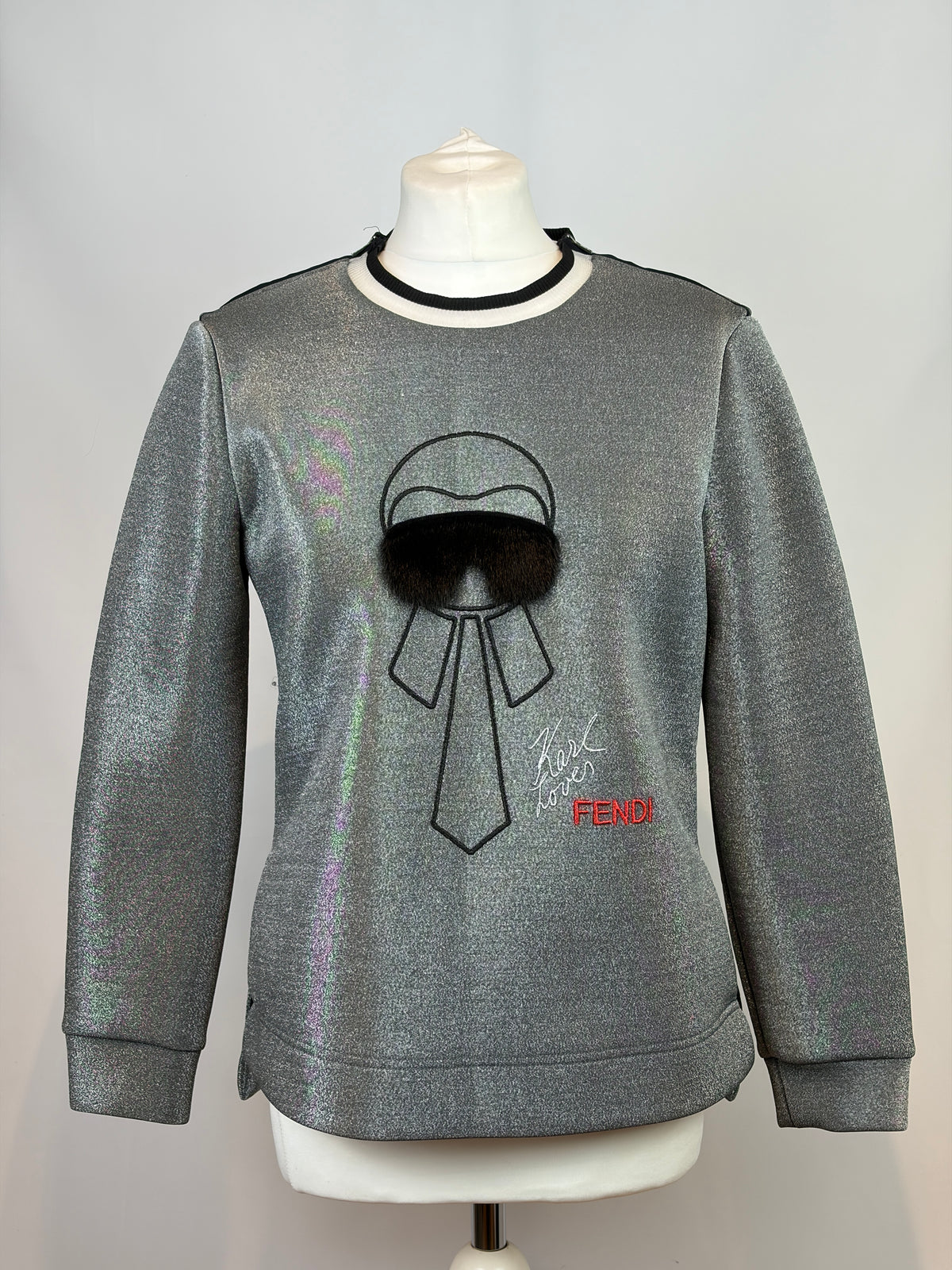 A metallic silver crewneck sweatshirt displayed on a white mannequin torso. The sweatshirt features a black embroidered bow and hat graphic with a black faux-fur pom-pom, a small red Fendi logo, and a black collar trim.