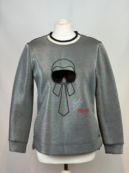 A metallic silver crewneck sweatshirt displayed on a white mannequin torso. The sweatshirt features a black embroidered bow and hat graphic with a black faux-fur pom-pom, a small red Fendi logo, and a black collar trim.