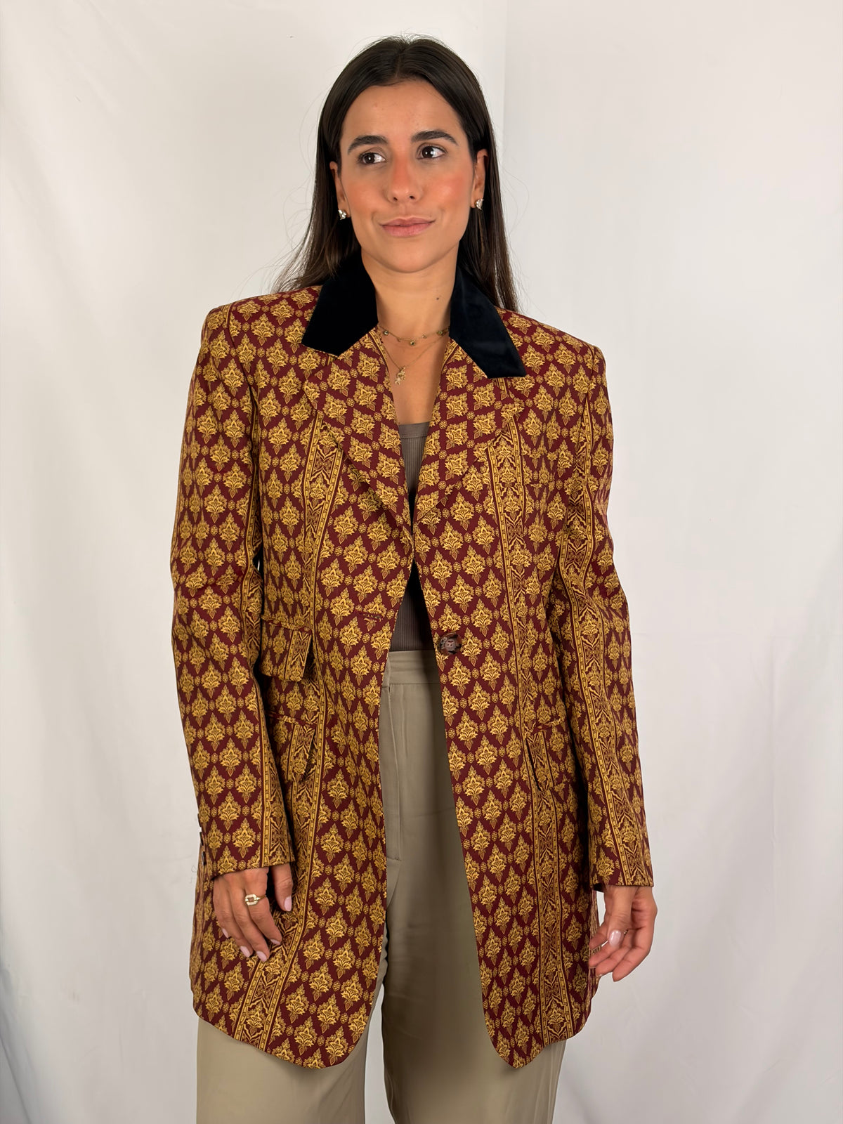 A woman modeling a vintage-style tailored blazer with a brown and gold geometric pattern and a black collar, paired with beige trousers, against a white studio backdrop.