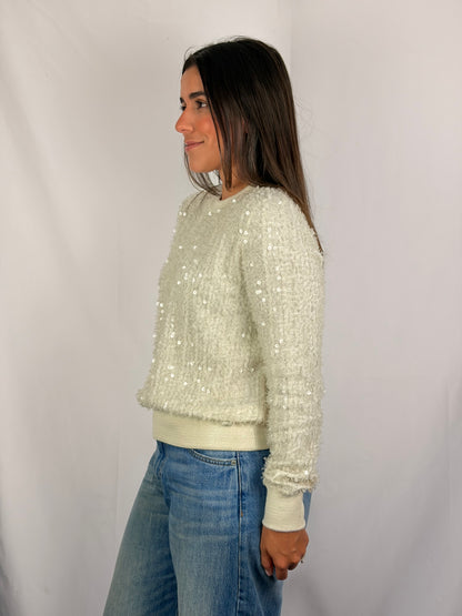 A woman wearing a cream-colored, long-sleeve knit sweater covered in small sequins, paired with blue jeans, standing against a white studio backdrop.
