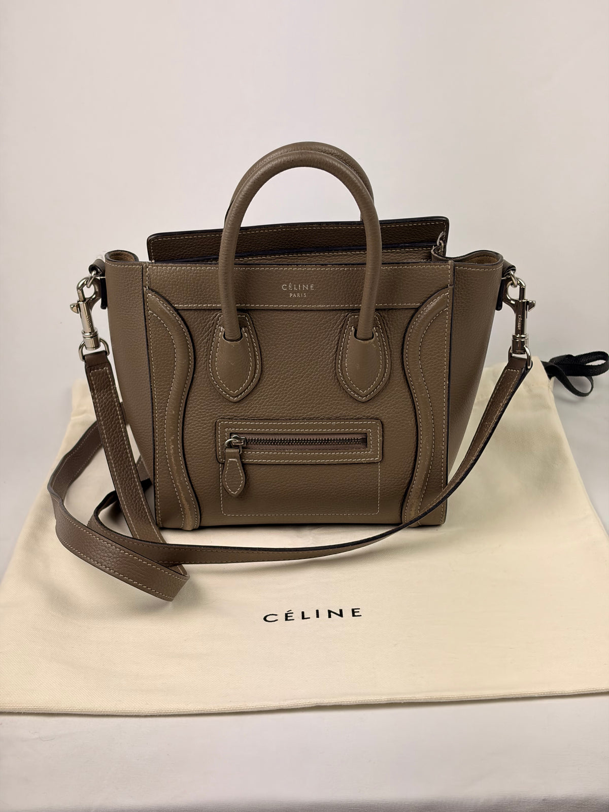 A taupe brown Celine Mini Luggage handbag with dual top handles and a detachable shoulder strap, displayed on a cream Celine dust bag against a light gray background.