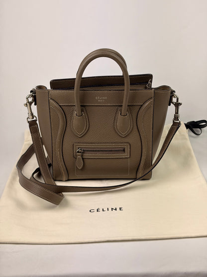 A taupe brown Celine Mini Luggage handbag with dual top handles and a detachable shoulder strap, displayed on a cream Celine dust bag against a light gray background.