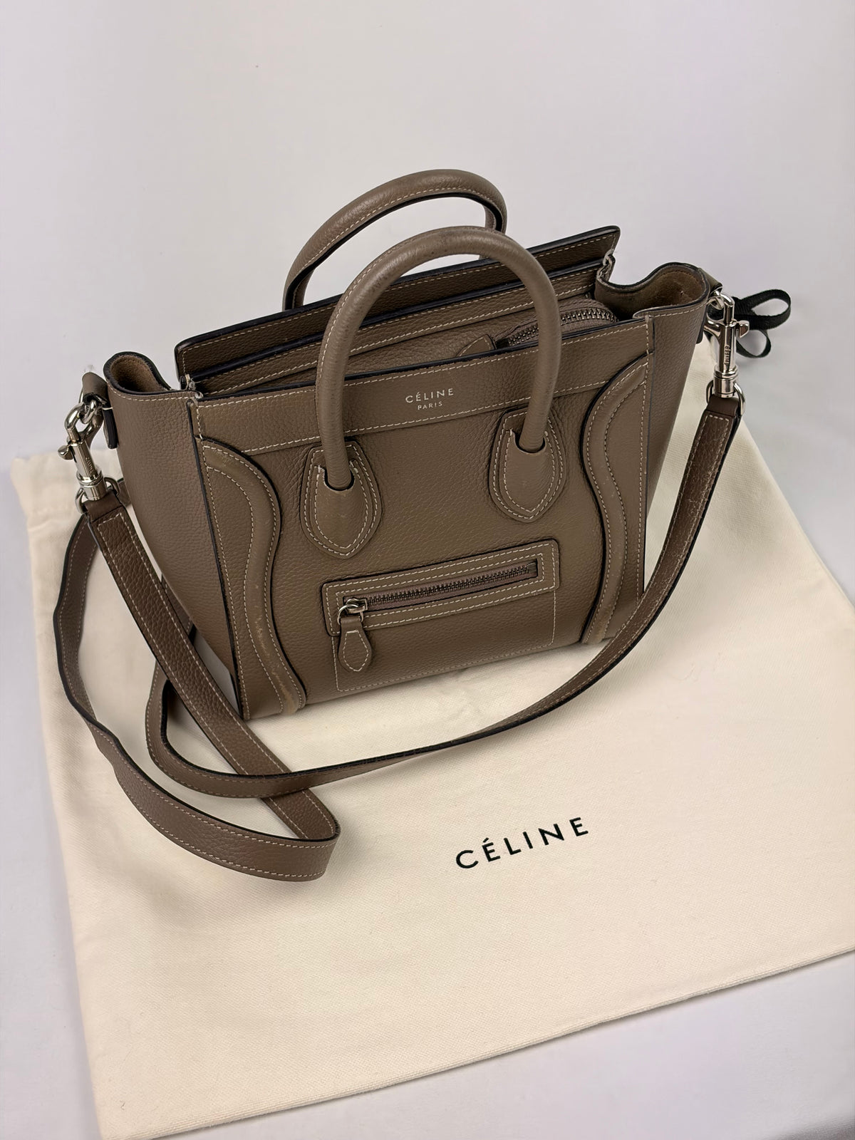 A taupe Céline handbag placed on top of a cream Céline dust bag against a plain light background. The handbag has dual top handles, a shoulder strap, and a front zip pocket.