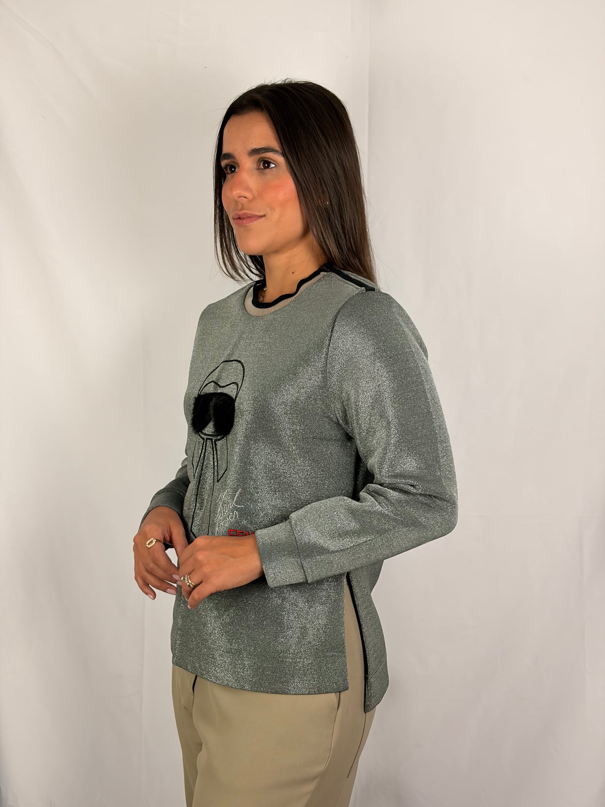 A woman modeling a long-sleeve metallic sage green top with black trim at the crew neck and a black graphic print on the front, paired with beige pants, against a white studio backdrop.