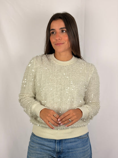 A woman modeling a cream-colored, long-sleeve crewneck sweater covered in small sequins, paired with blue jeans, against a plain white studio background.