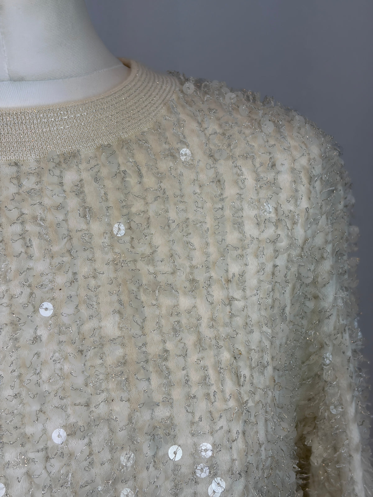 Close-up of a cream-colored textured knit sweater with sequins, displayed on a mannequin torso against a neutral gray background.