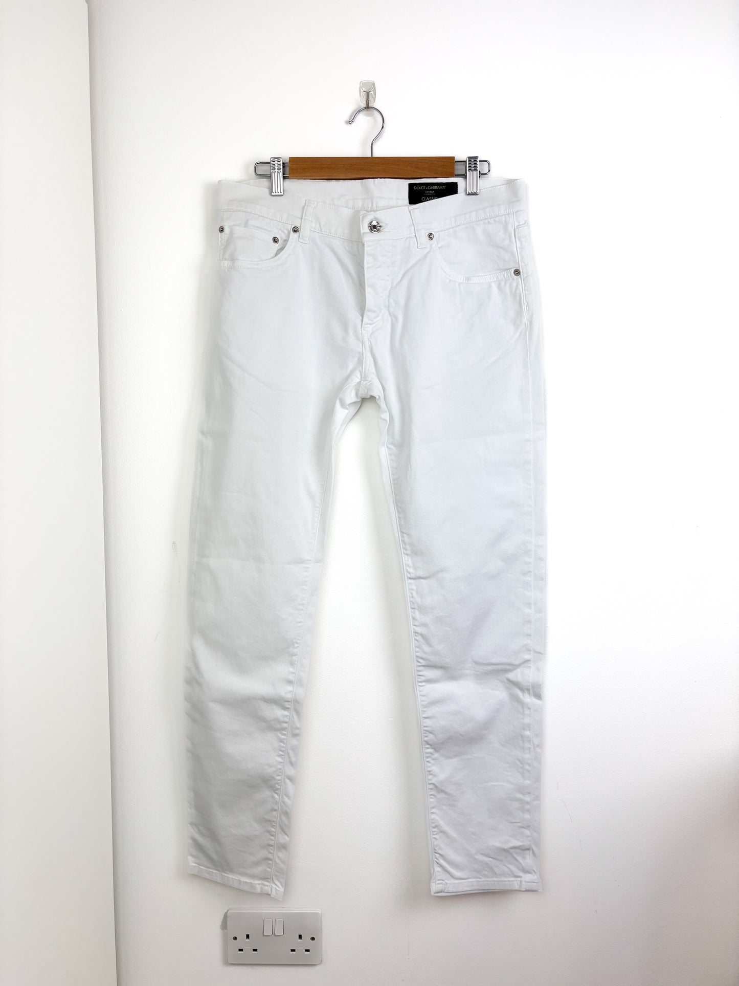 White jeans hanging on a wooden hanger against a white wall.