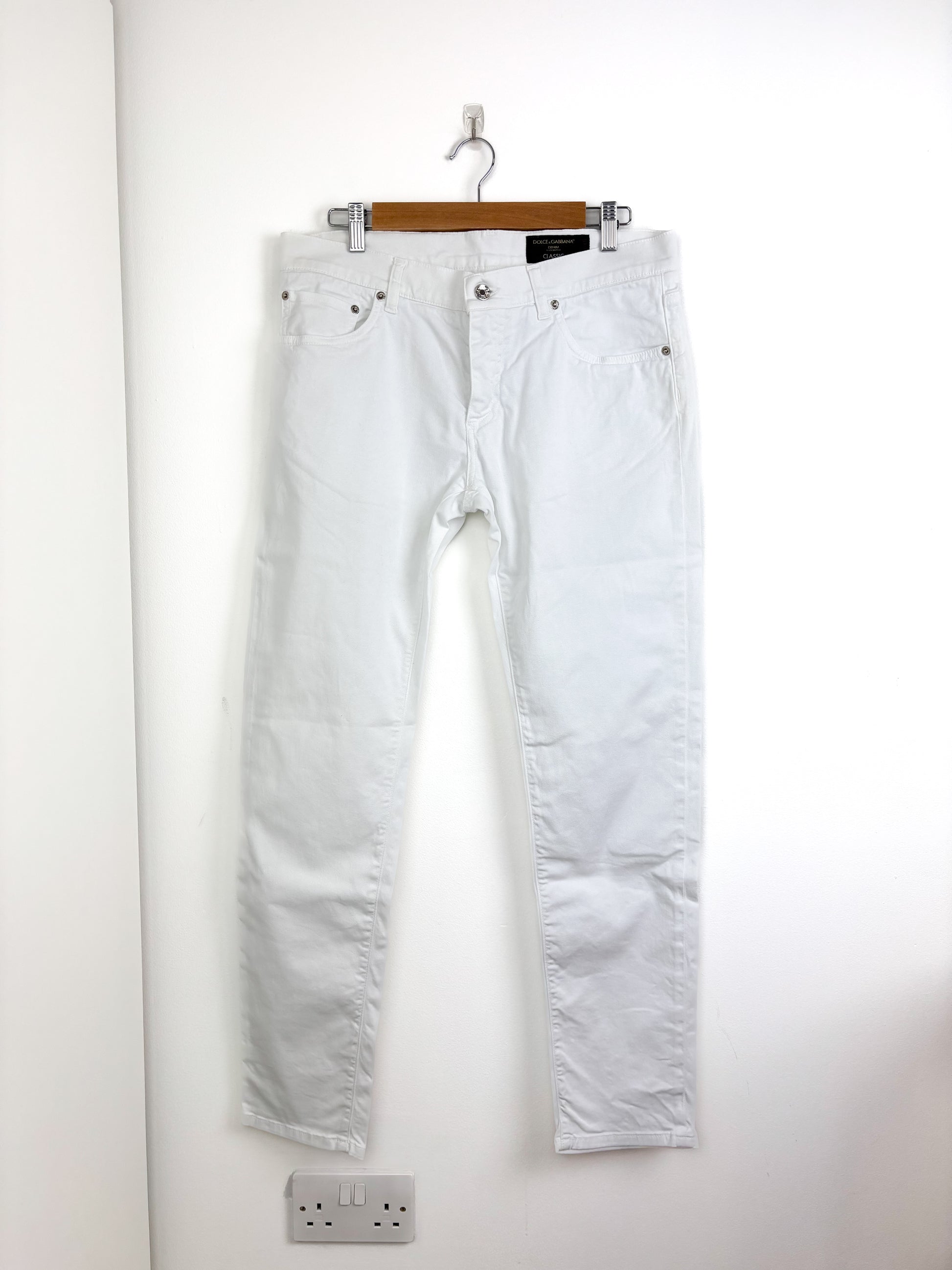 White jeans hanging on a wooden hanger against a white wall.